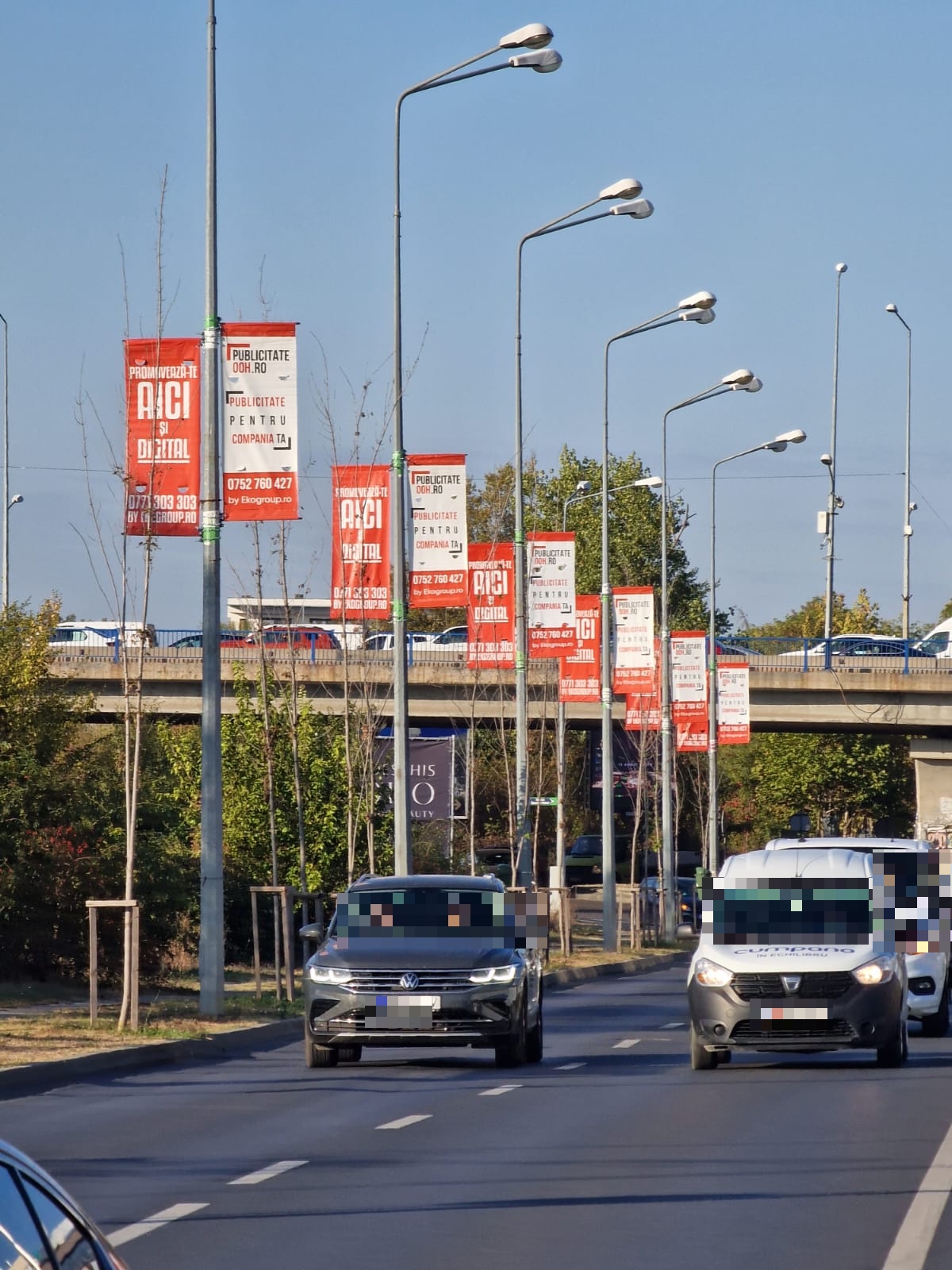 Trafic auto pe o arteră urbană, cu steaguri direcționale montate în perechi pe stâlpii de iluminat de pe marginea drumului